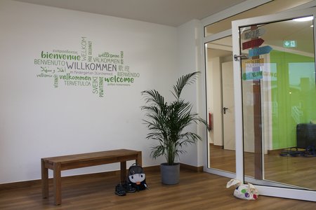 Eingangshalle der Kita Storchennest in Treia. Auf der weißen Wand stehen in grün Willkommensgrüße in verschiedenen Sprachen. 