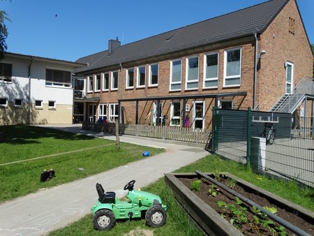 Außenbereich der Kita Hollerbue. Vorne im Bild ein Spielfahrzeug und ein Blumenbeet. 